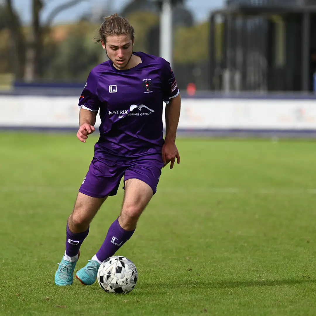 Male soccer player in a purple kit dribbling the ball on a grassy field during a match.